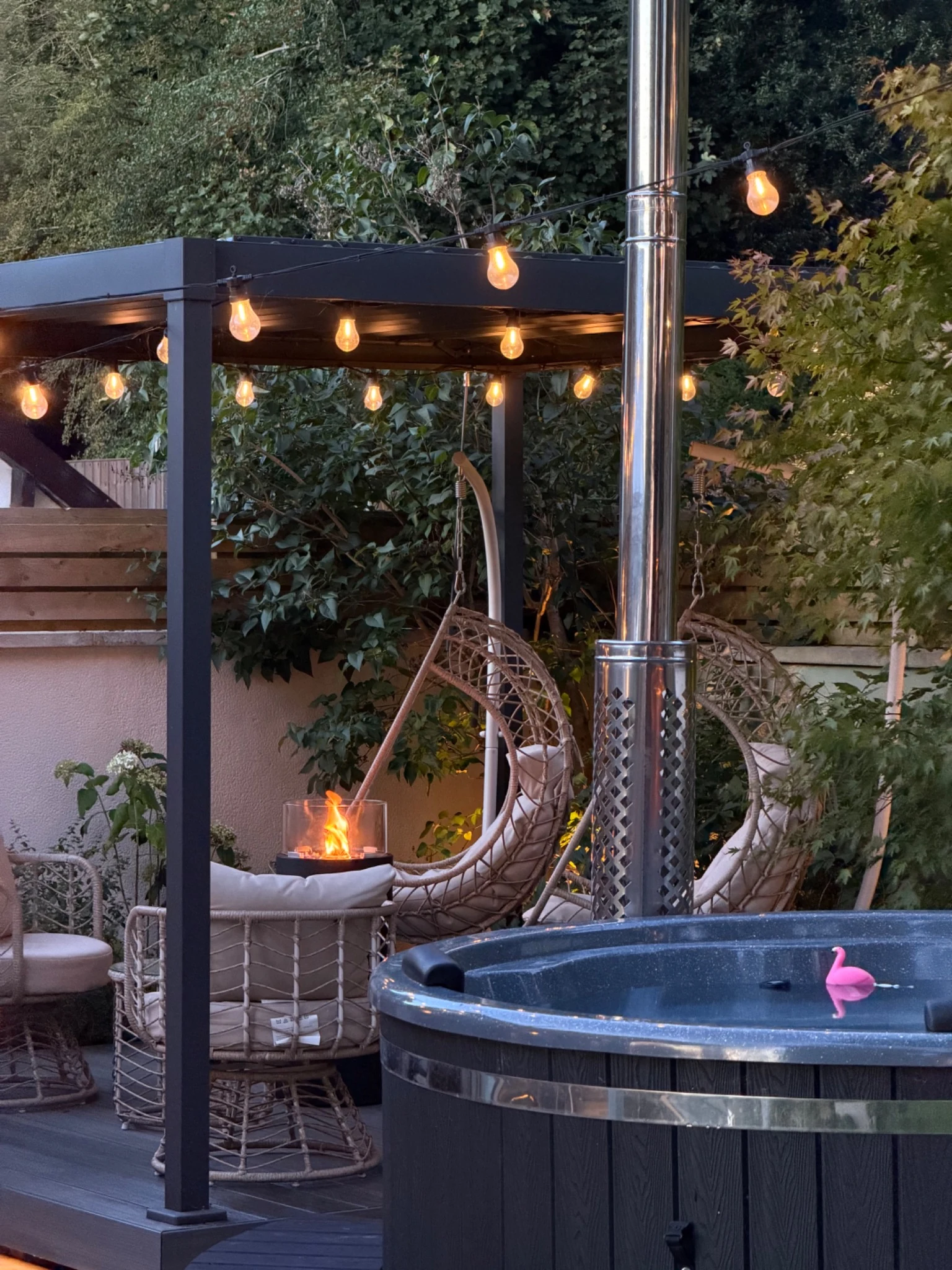 Wood-fired hot tub beside a sheltered pergola with festoon lighting and garden seating